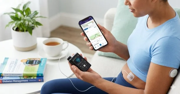 A side view of a patient adjusting a computerized insulin pump device, displaying the connection tubing attached to a subcutaneous abdominal infusion set alongside a smartphone trend dashboard.